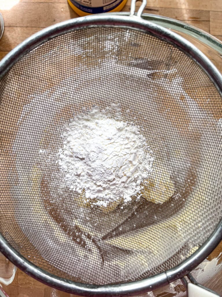 Icing sugar being sifted into whipped butter in the bowl of an electric stand mixer.