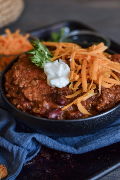 A bowl of chili topped with shredded cheese and sour cream, on a tray with a blue napkin.