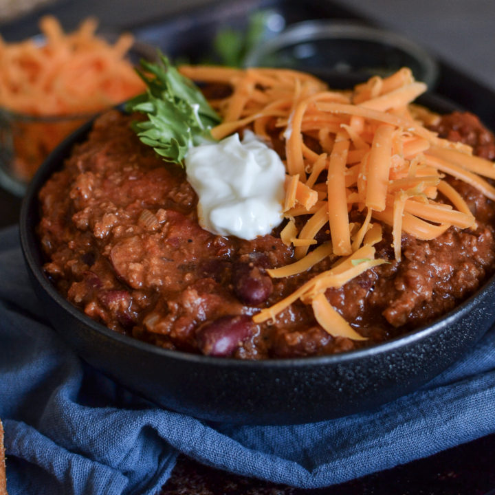 A bowl of chili topped with shredded cheese and sour cream, on a tray with a blue napkin.