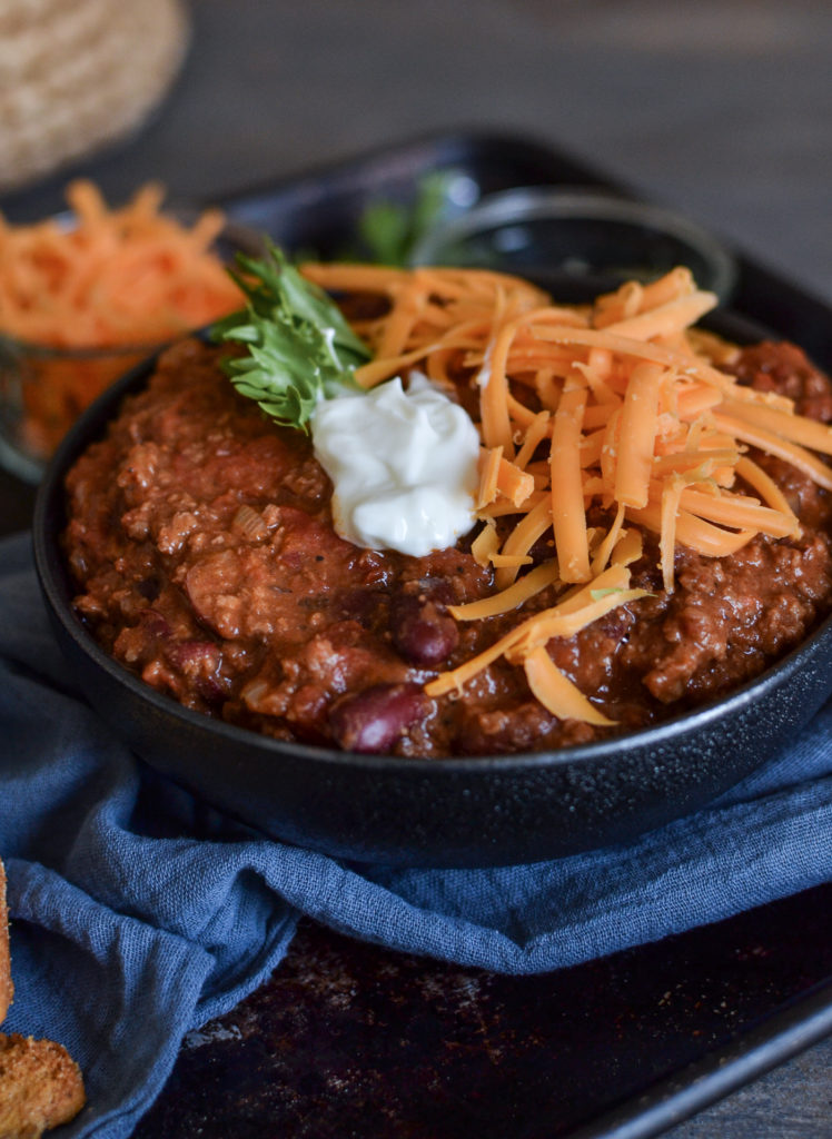 A bowl of chili topped with shredded cheese and sour cream, on a tray with a blue napkin.