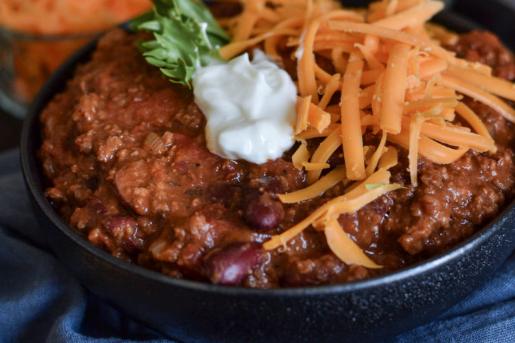 A bowl of chili topped with shredded cheese and sour cream, on a tray with a blue napkin.