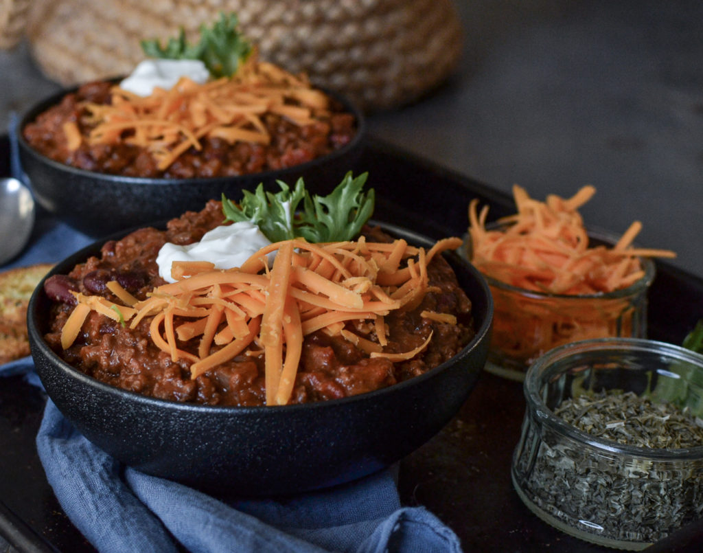 Two bowls of chili topped with grated cheddar cheese and sour cream, set on a denim blue napkin.