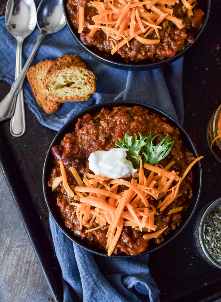 Overhead view of a bowl of chili topped with shredded cheddar cheese, a dollop of sour cream and parsley with a side of mini garlic toasts.