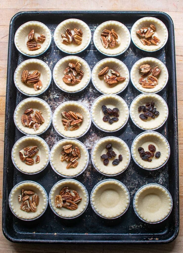 Tart shells on a baking sheet, most filled with pecans, 4 filled with raisins and 2 left empty.
