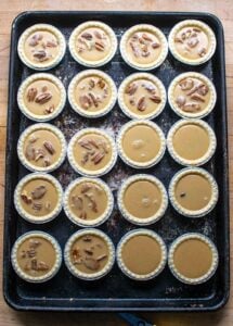 Tart shells on a baking sheet, most filled with pecans and butter tart filling, 4 filled with raisins and butter tart filling and 2 with just butter tart filling.
