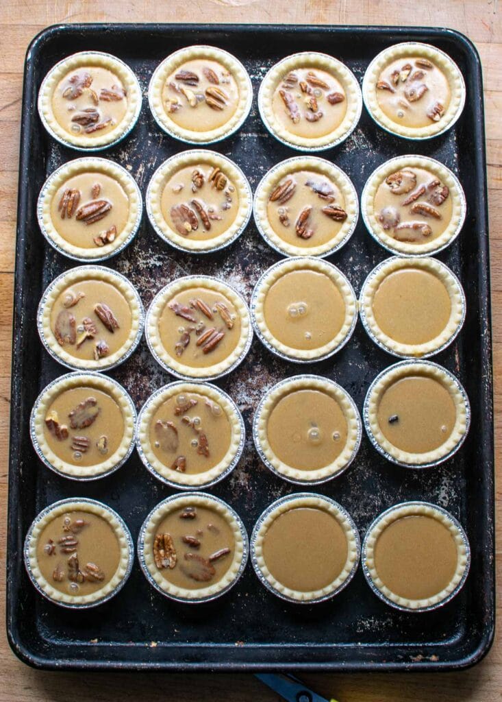 Tart shells on a baking sheet, most filled with pecans and butter tart filling, 4 filled with raisins and butter tart filling and 2 with just butter tart filling.
