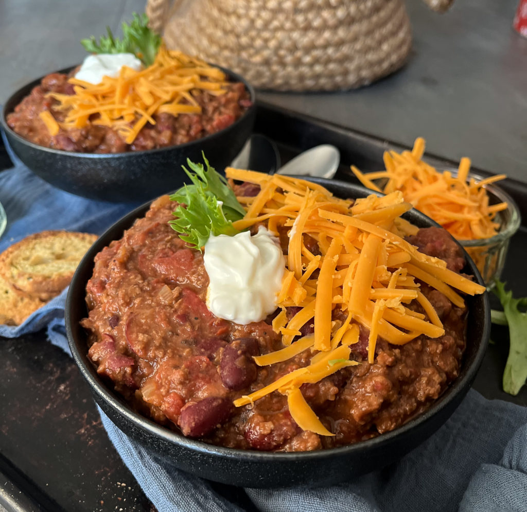 Two bowls of chili topped with shredded cheddar cheese, a dollop of sour green and a spring of parsley.