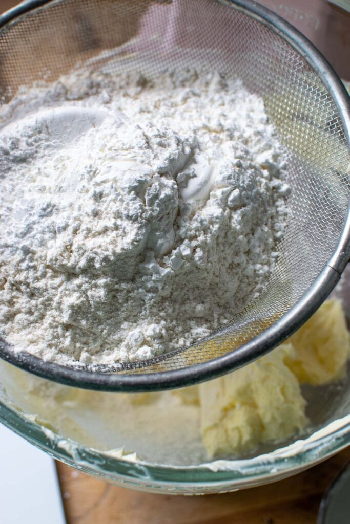 A mixture of flour, powdered sugar, and cornstarch being sifted into a mixing bowl of whipped butter.