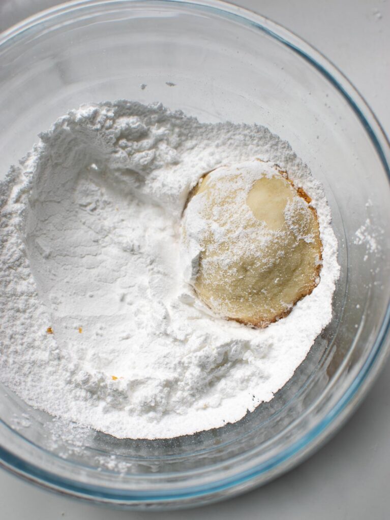 A warm shortbread snowball cookie being rolled in a bowl of confectioners sugar.