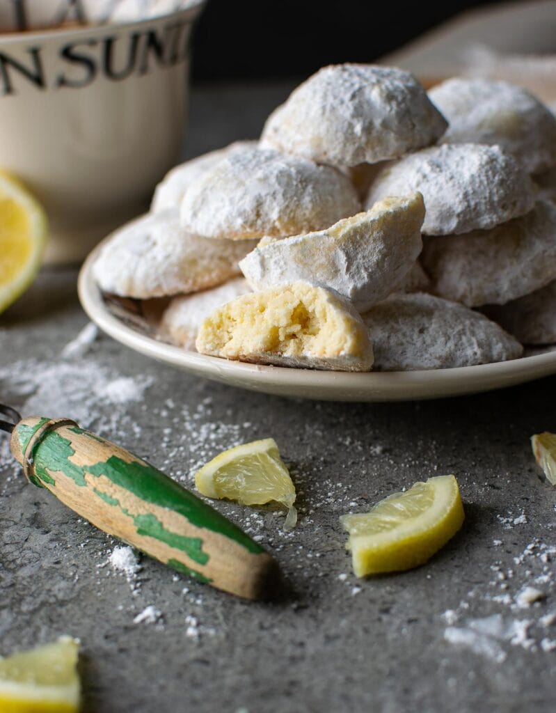 Lemon snowball cookies piled on a plate, with one cut in half to show the inside.  Little sections of lemon surround the plate.