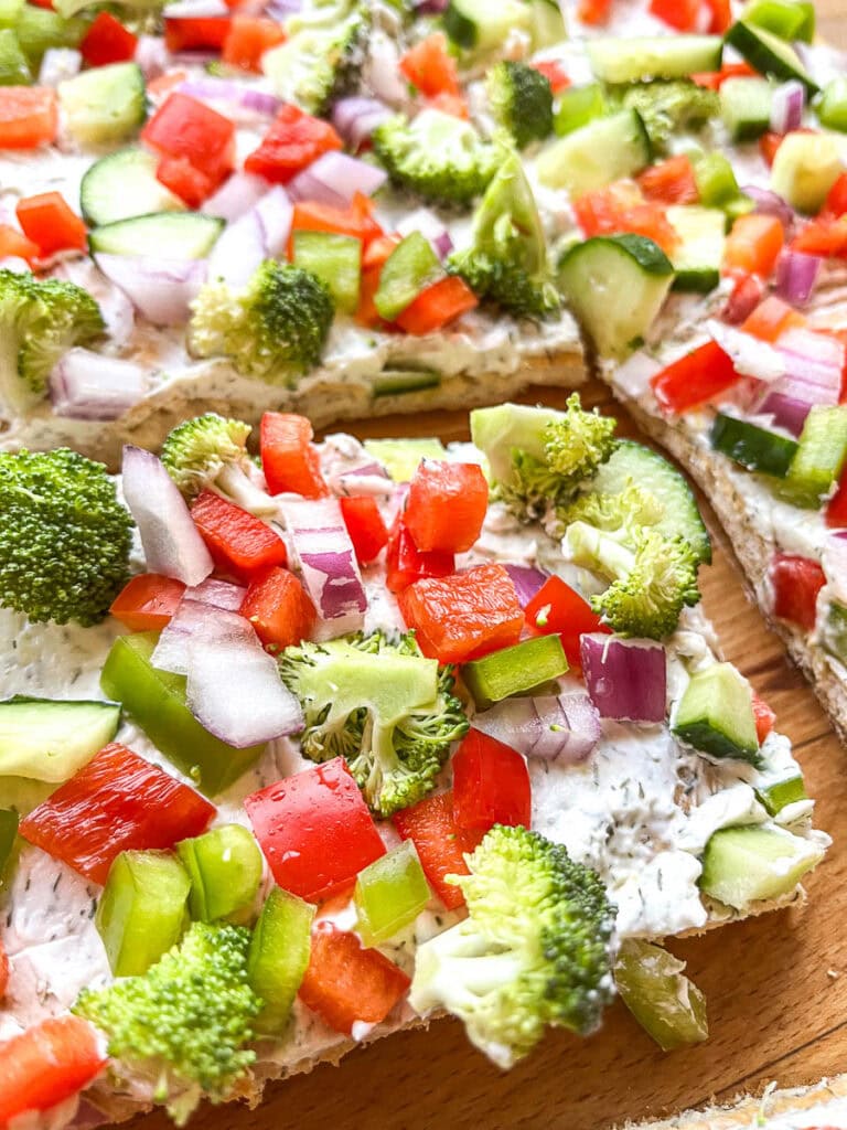A close up of veggie cream cheese squares topped with fresh red peppers, cucumbers, red onion and broccoli.