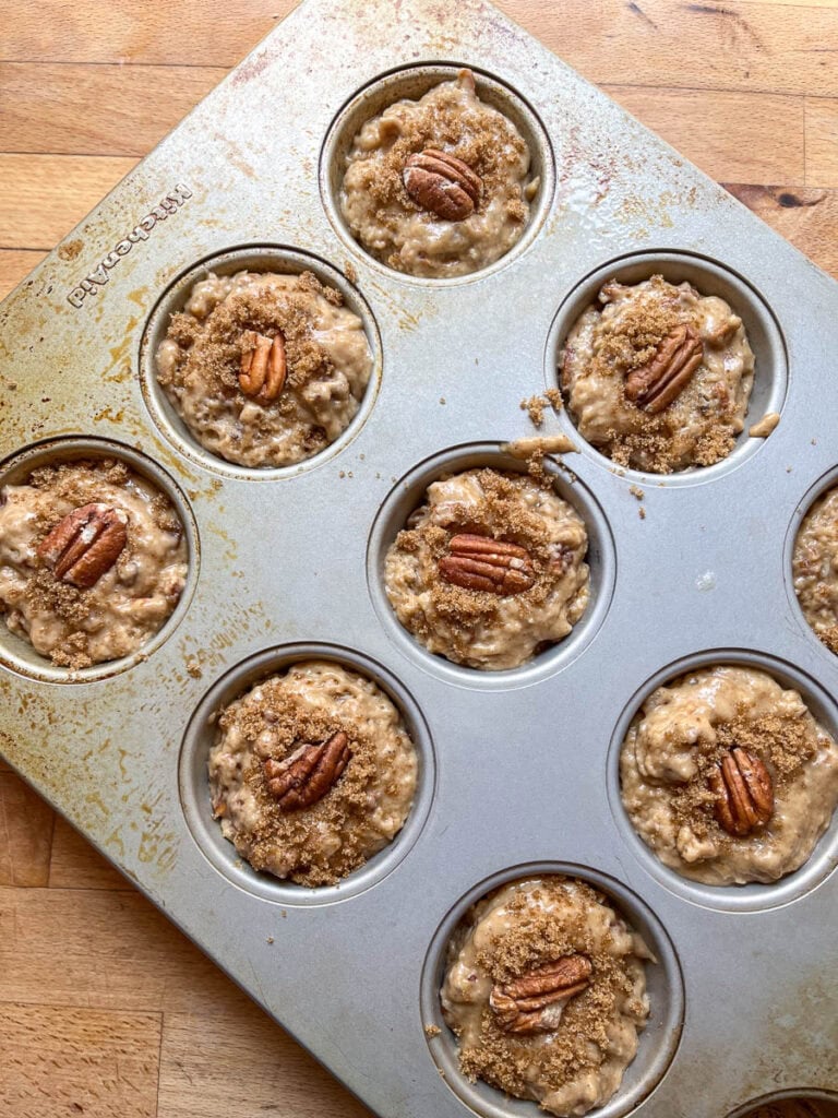 Butter pecan muffin batter in muffin tins and topped with brown sugar crumble and a pecan half, before baking.