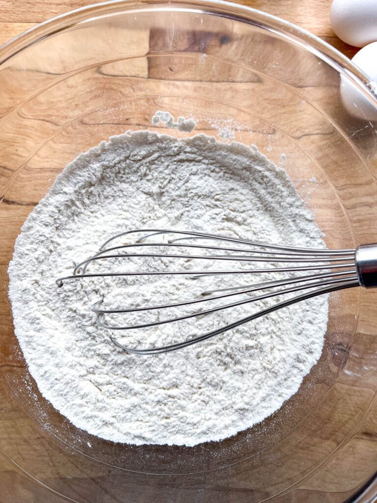 Flour, baking soda, baking powder and salt whisked together in a large glass bowl, with the whisk still in the bowl.