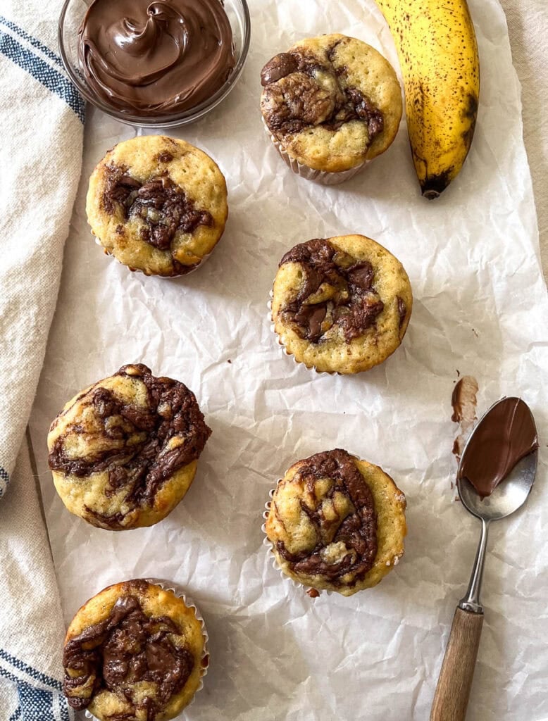 Nutella Banana muffins on a piece of parchment paper with an over ripe banana and a small bowl of Nutella beside.
