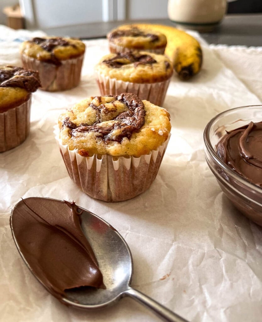 Banana Nutella Muffins on a piece of wrinkled parchment paper with a spoonful and bowl of Nutella to the side.