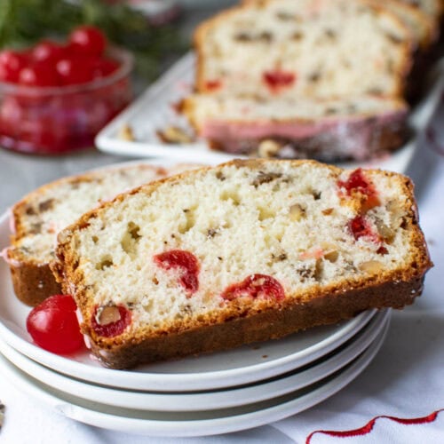 Slices of cherry nut bread on a stack of white plates and a small bowl of maraschino cherries in the background.