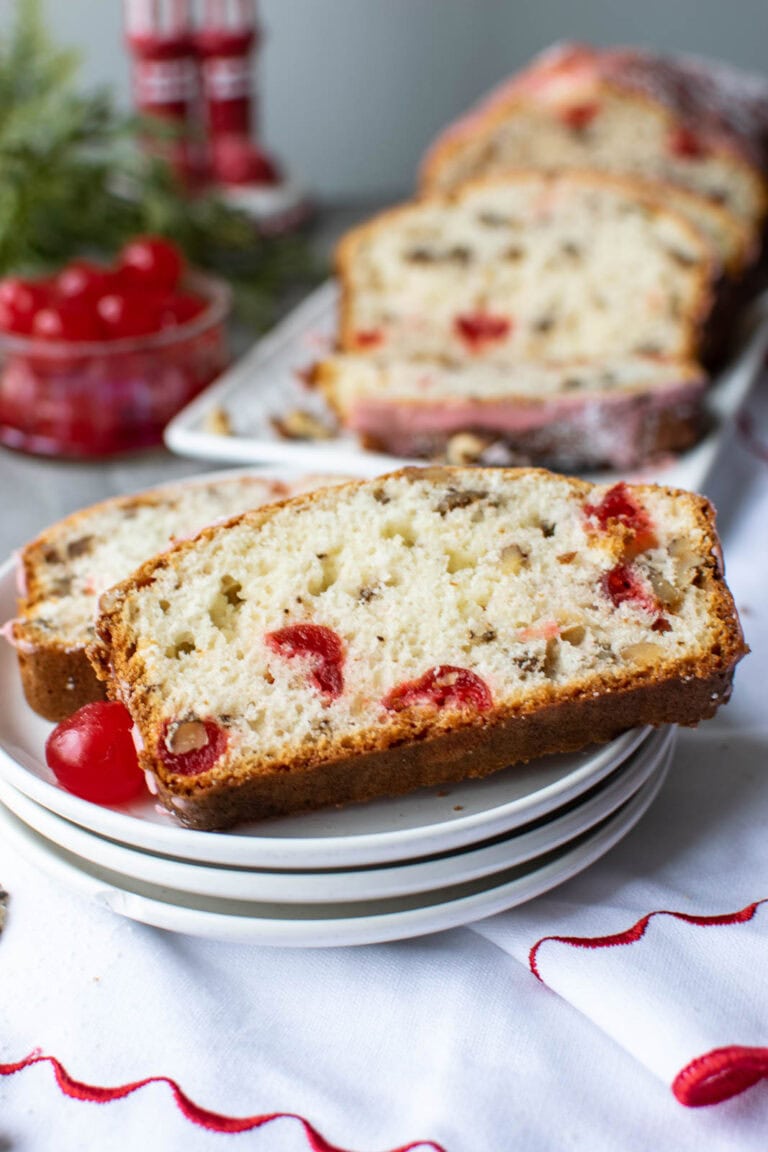 Slices of cherry nut bread on a stack of white plates and a small bowl of maraschino cherries in the background.