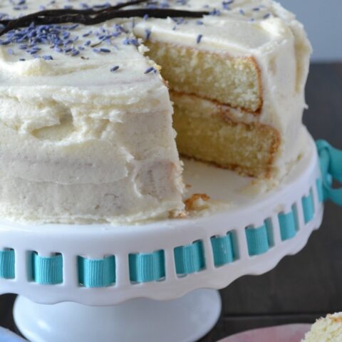 A vanilla lavender cake with a slice cut out of it on a white glass cake stand with blue ribbon.