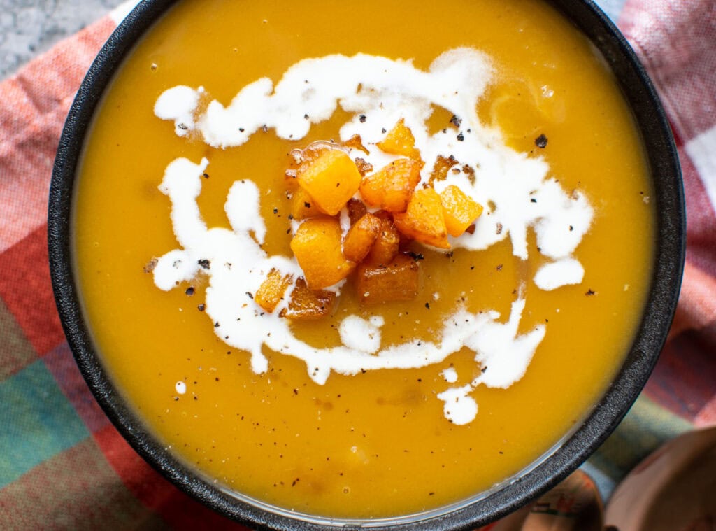 Close up view of a bowl of butternut squash soup topped with bits of pan fried squash, a swirl of cream and maple syrup.