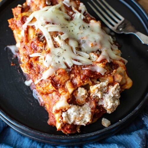 3 Cheese manicotti on a black dinner plate sitting on top of a blue napkin, with a fork to the side.