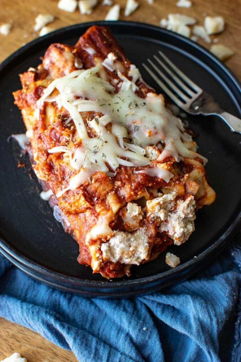 3 Cheese manicotti on a black dinner plate sitting on top of a blue napkin, with a fork to the side.