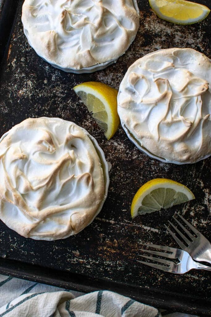 2 lemon meringue tarts made without crusts in ramekin dishes, with slices of lemon and forks on a baking tray.