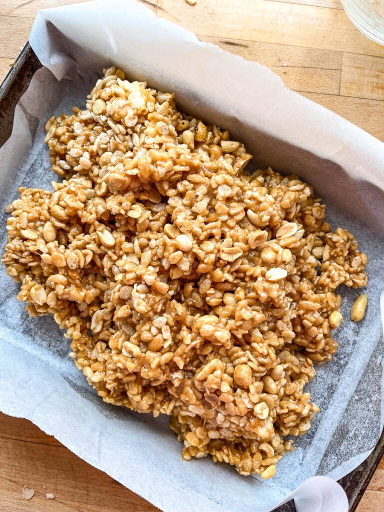 Mixture of Rice Krispie cereal, peanuts and a creamy peanut butter base in a parchment lined 9 by 13 inch pan before pressing it down.