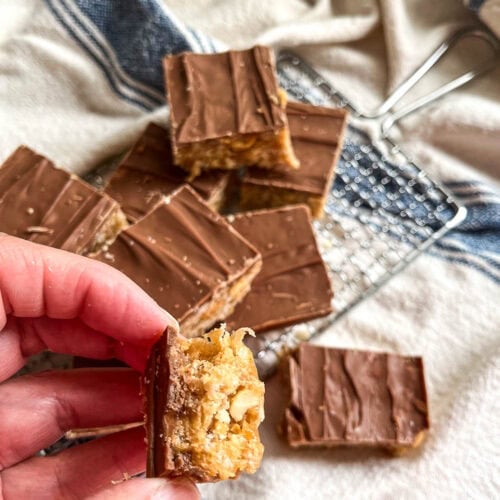 Squares of chocolate topped crispy bars on a small cooling rack with one square being held by a hand.