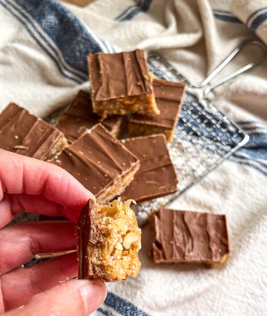 Squares of chocolate topped crispy bars on a small cooling rack with one square being held by a hand.