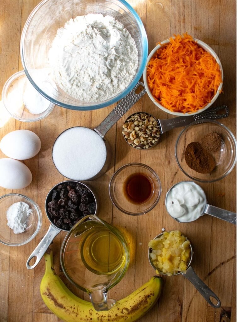 All the ingredients to make morning glory muffins including shredded carrot, crushed pineapple, mashed banana, eggs, flour, sugar and spices.