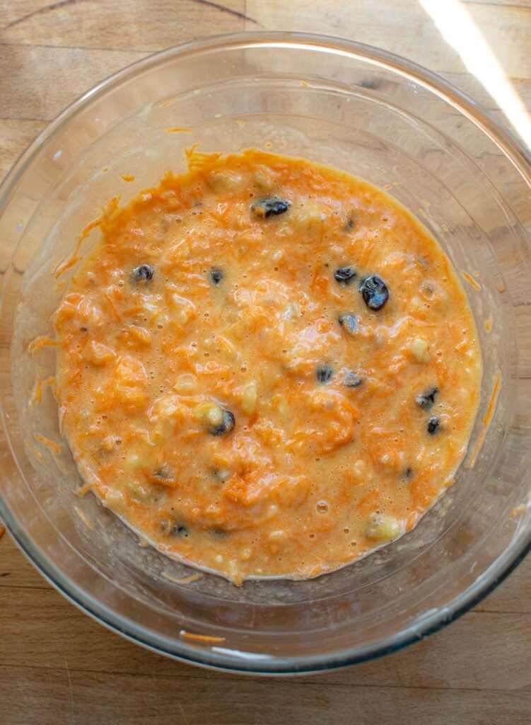 Shredded carrots, crushed pineapple, mashed banana, raisins and yogurt stirred into egg mixture in a glass mixing bowl.