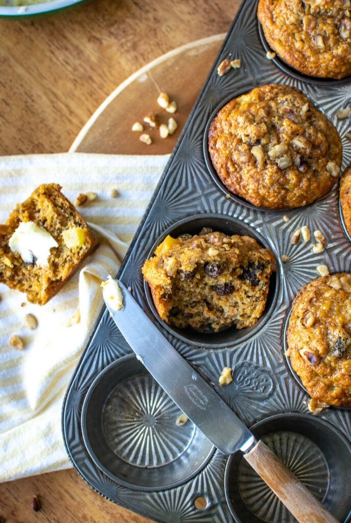 This image shows the corner of a muffin pan with 3 visible muffins, a butter knife across the pan and one muffin out of the pan, torn in half with butter on it.
