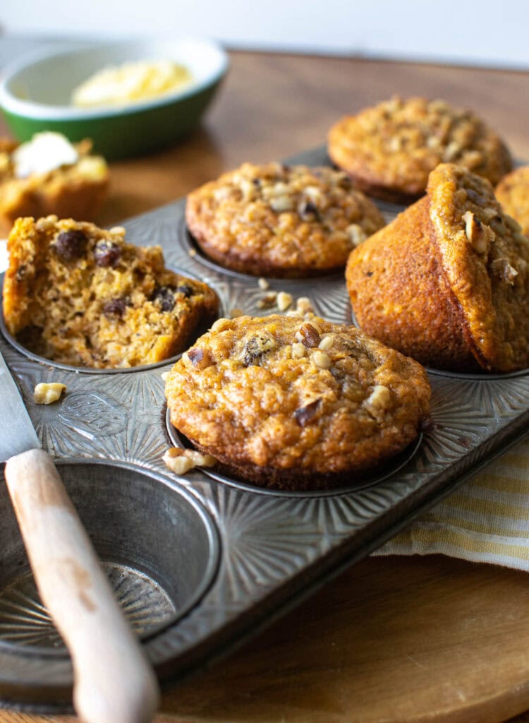 A muffin tray with freshly baked morning glory muffins, one propped up out of the tin and one torn in half.