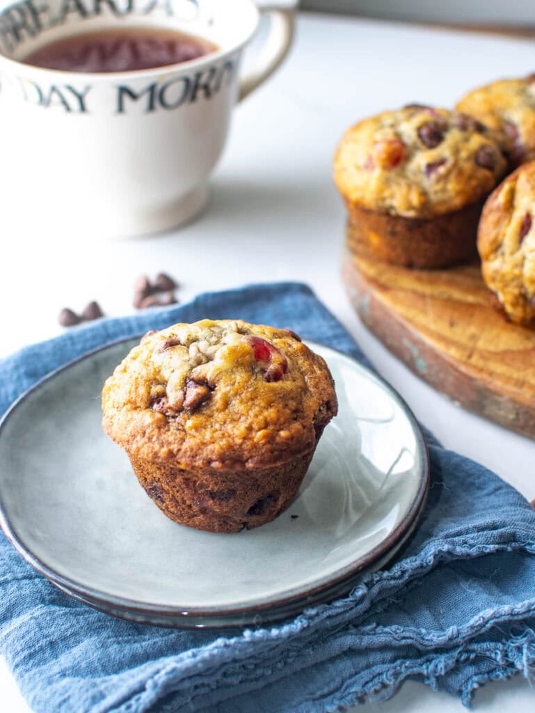 A cherry chocolate chip muffin on a plate with a cup of tea in the background, and more muffins on a cutting board.