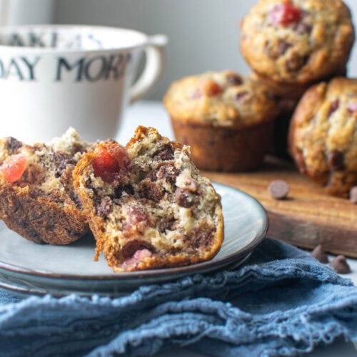 A homemade cherry chocolate muffin split in half on a plate with 3 more muffins in the background.