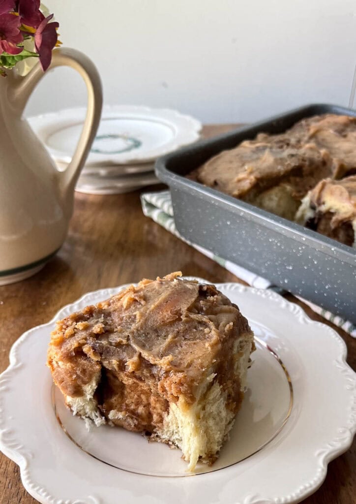 A pecan cinnamon roll on a white plate in the foreground, with a pan of cinnamon rolls in the background.