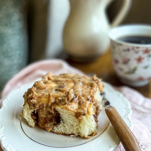 A pecan cinnamon roll on a white plate with a cup of coffee I the background.