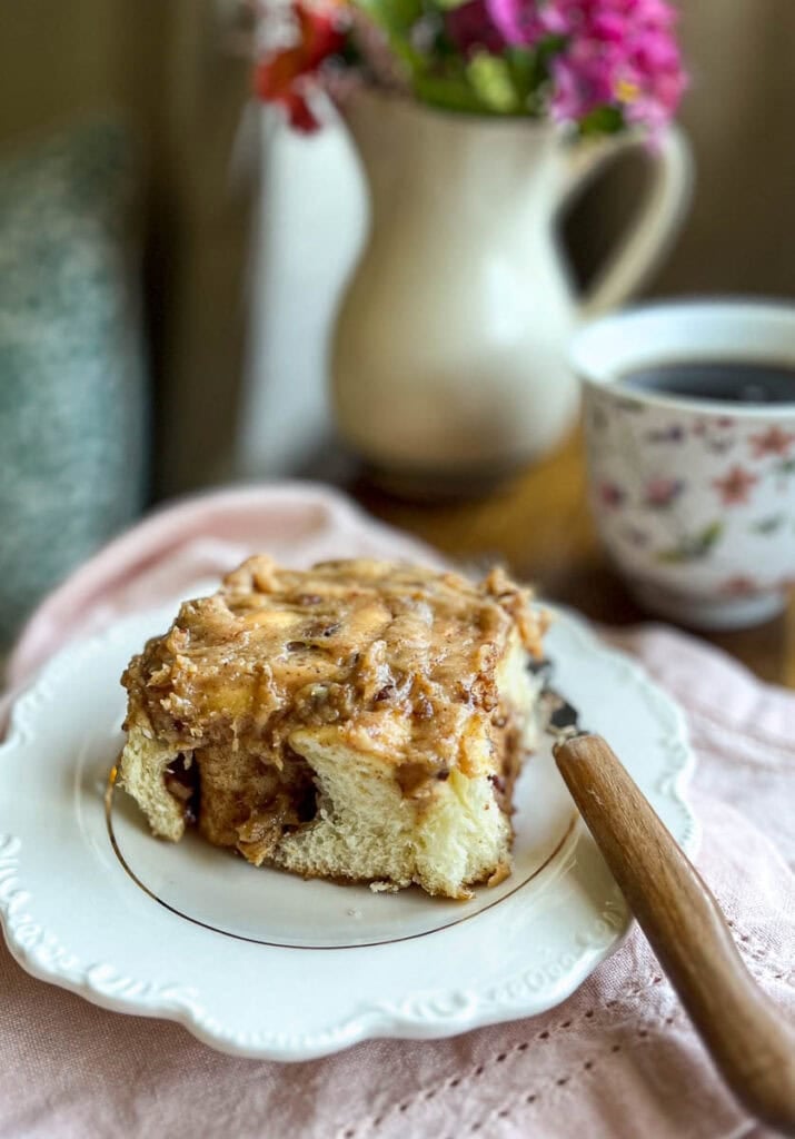 A pecan cinnamon roll on a white plate with a cup of coffee I the background.