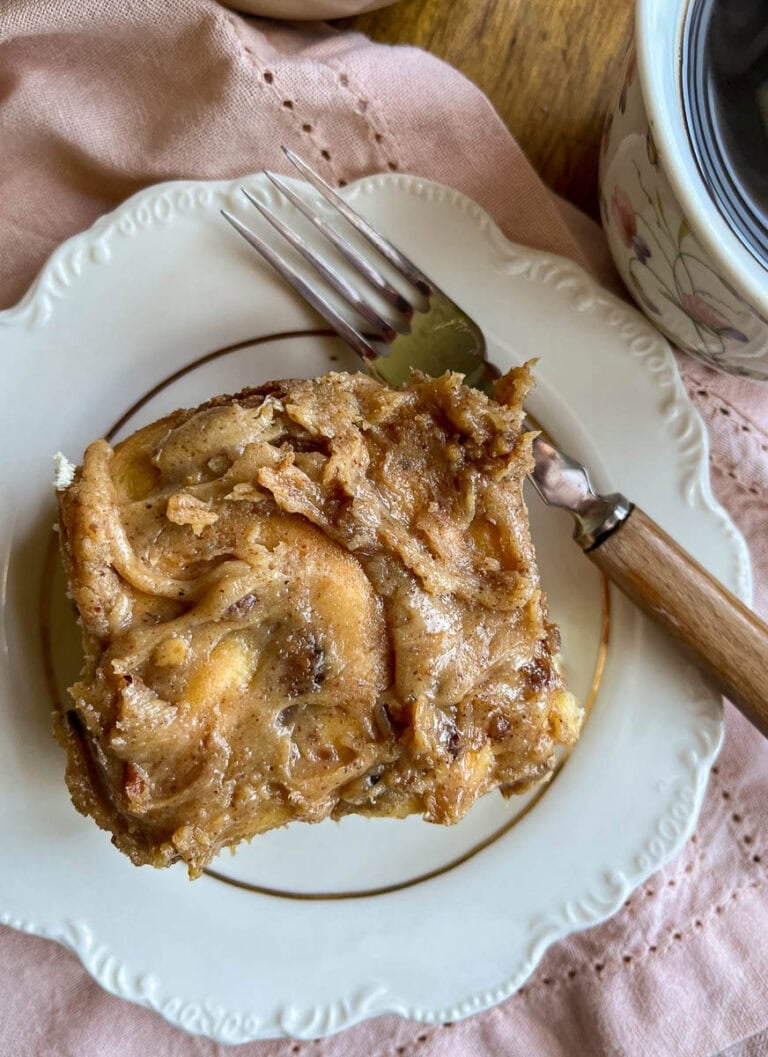 A pecan cinnamon roll frosted with pecan cream cheese frosting on a white plate with a fork beside it.