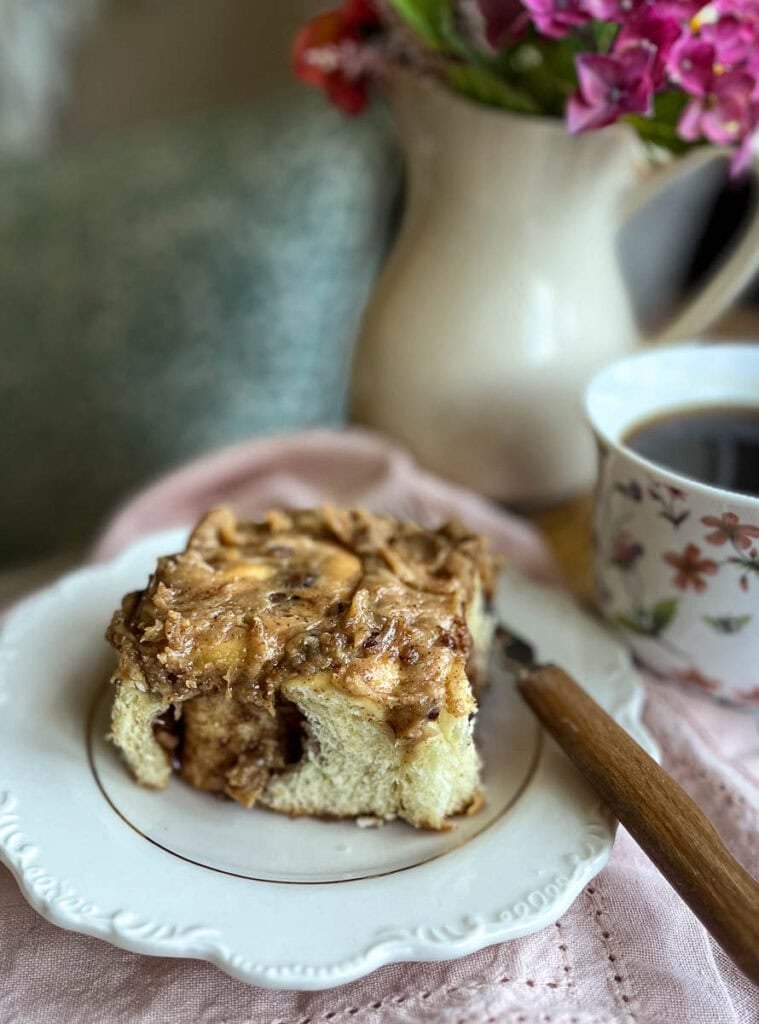 A pecan cinnamon roll on a plate beside a cup of coffee and a vase of flowers.