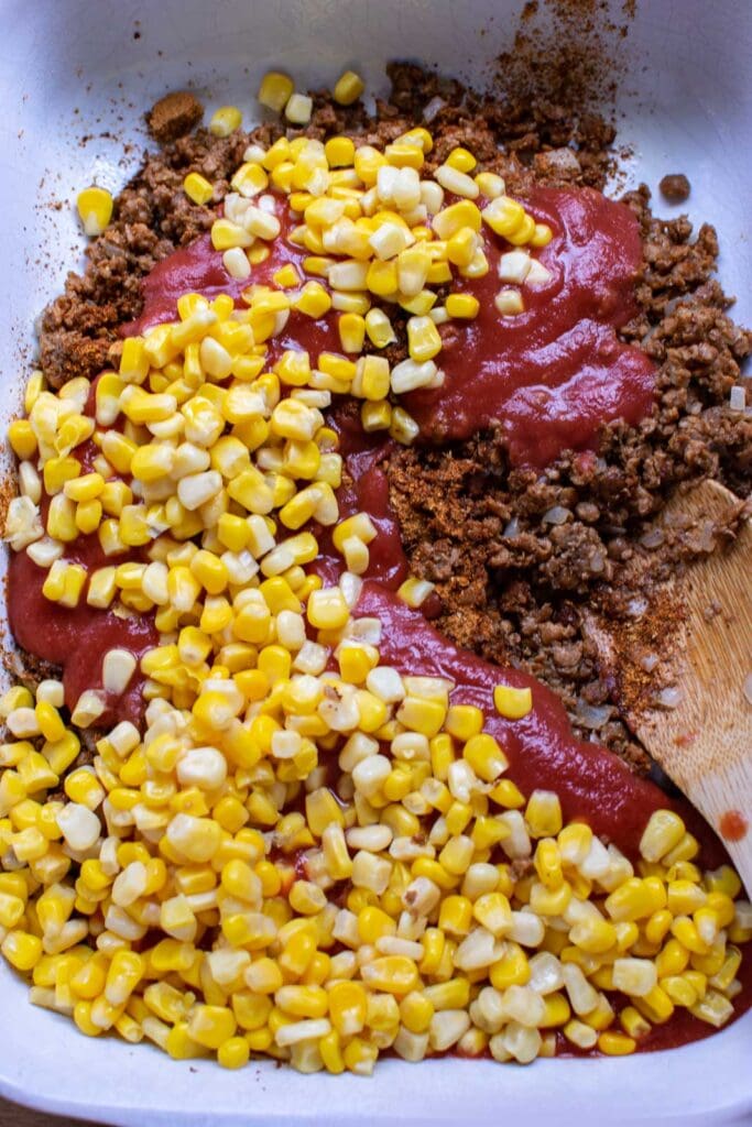 Browned ground beef and onion being mixed with tomato sauce, taco seasoning and corn in a casserole dish.