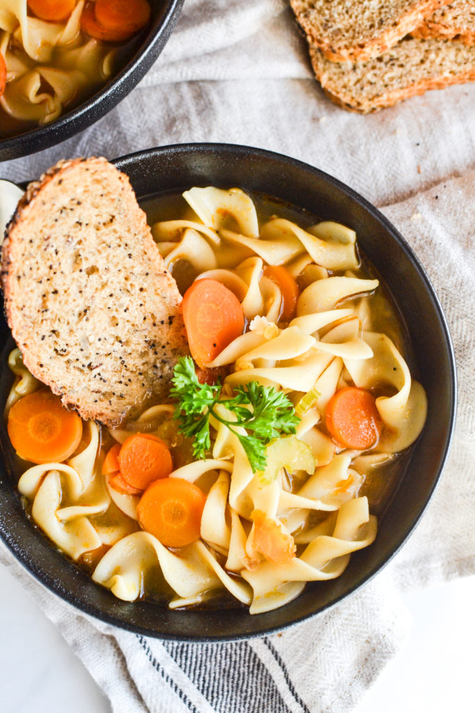 Black ceramic bowl filled with vegetable noodle soup and a wedge of sourdough bread