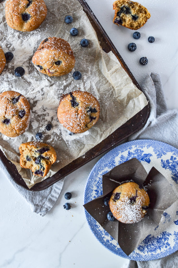 Overhead view of a pan of blueberry muffins with a single muffin on a blue transfer ware plate