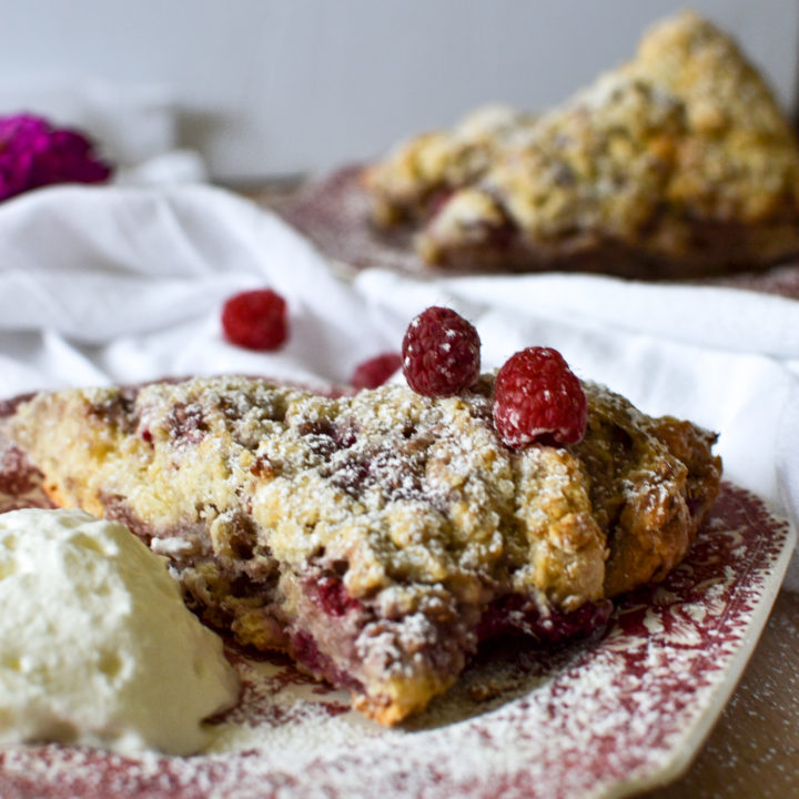 A white chocolate raspberry scone sprinkled with icing sugar and fresh raspberries with clotted cream beside