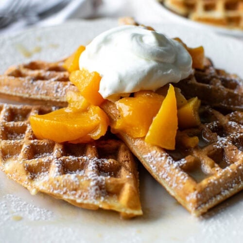 Crispy waffles topped with fresh peach sauce and whipped cream on a white serving plate.
