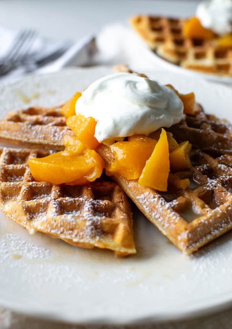 Crispy waffles topped with fresh peach sauce and whipped cream on a white serving plate.