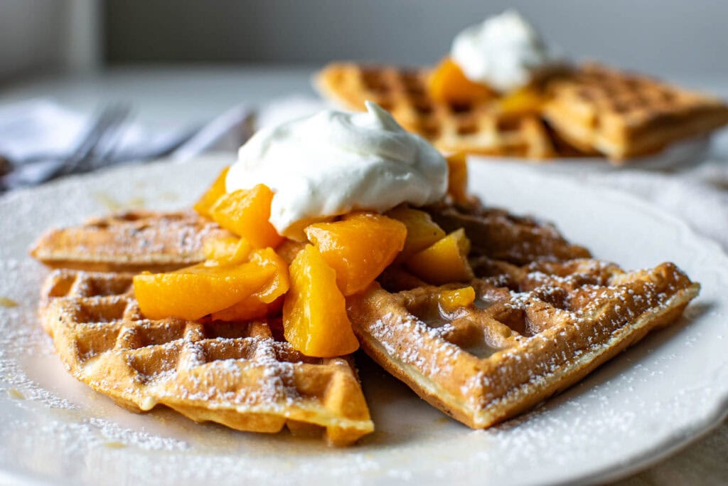Peach waffles with a peach sauce topping and a dollop of whipped cream served on a white plate.