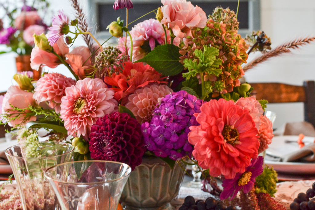 Dahlias and zinnias in orange, purple and pink arranged in a turquoise footed bowl.