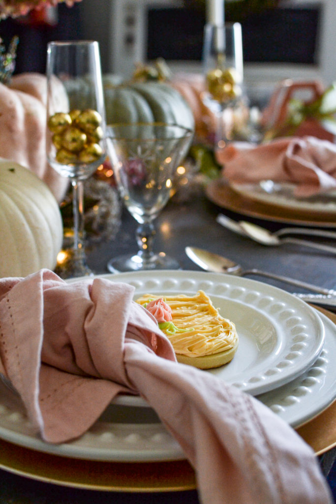 A table setting with white plates, a pumpkin sugar cookie, silverware and gold rimmed water glasses