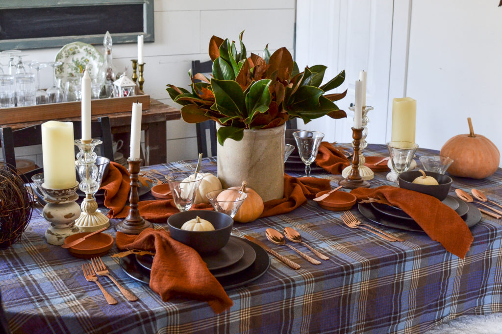 Thanksgiving tablescape with blue plaid tablecloth, magnolia leaf centerpiece, brown plates and terracotta napkins.